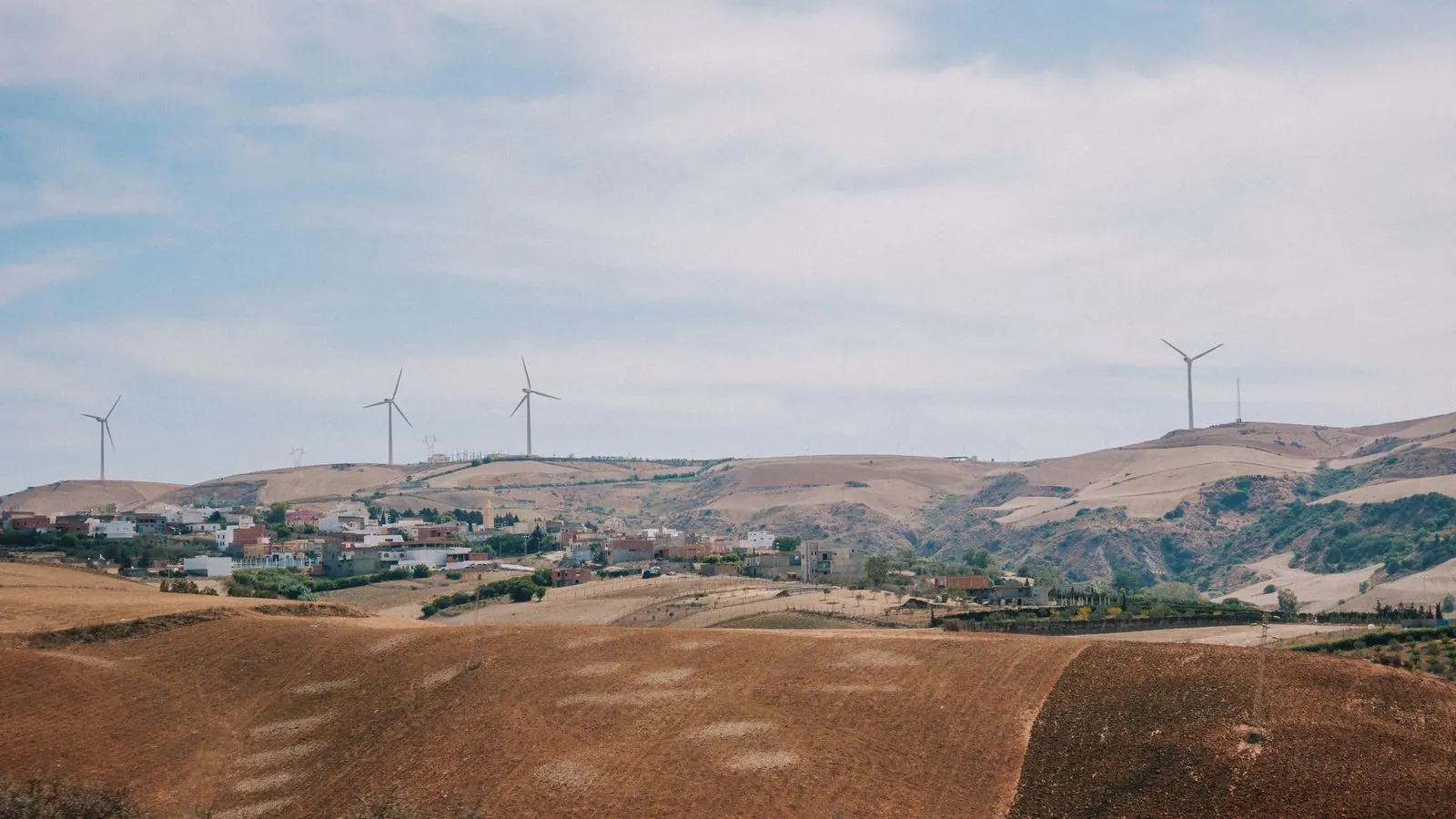 Panoramic view of a rural landscape with wind turbines on rolling hills and scattered buildings.