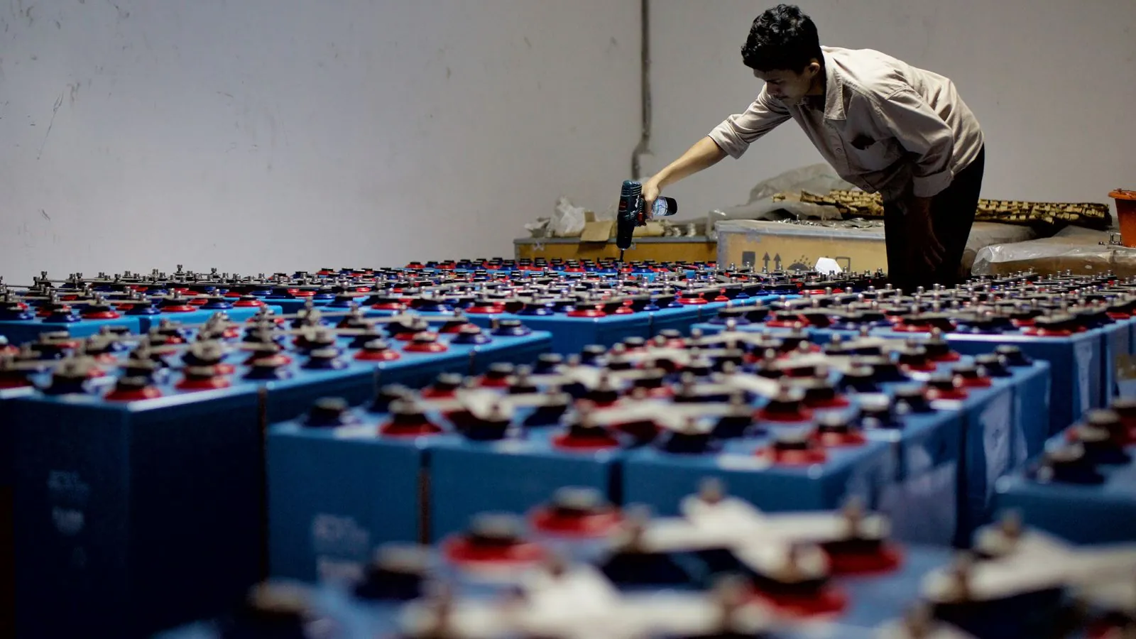 A worker checking many industrial batteries inside a facility. Indoor, industrial setting.
