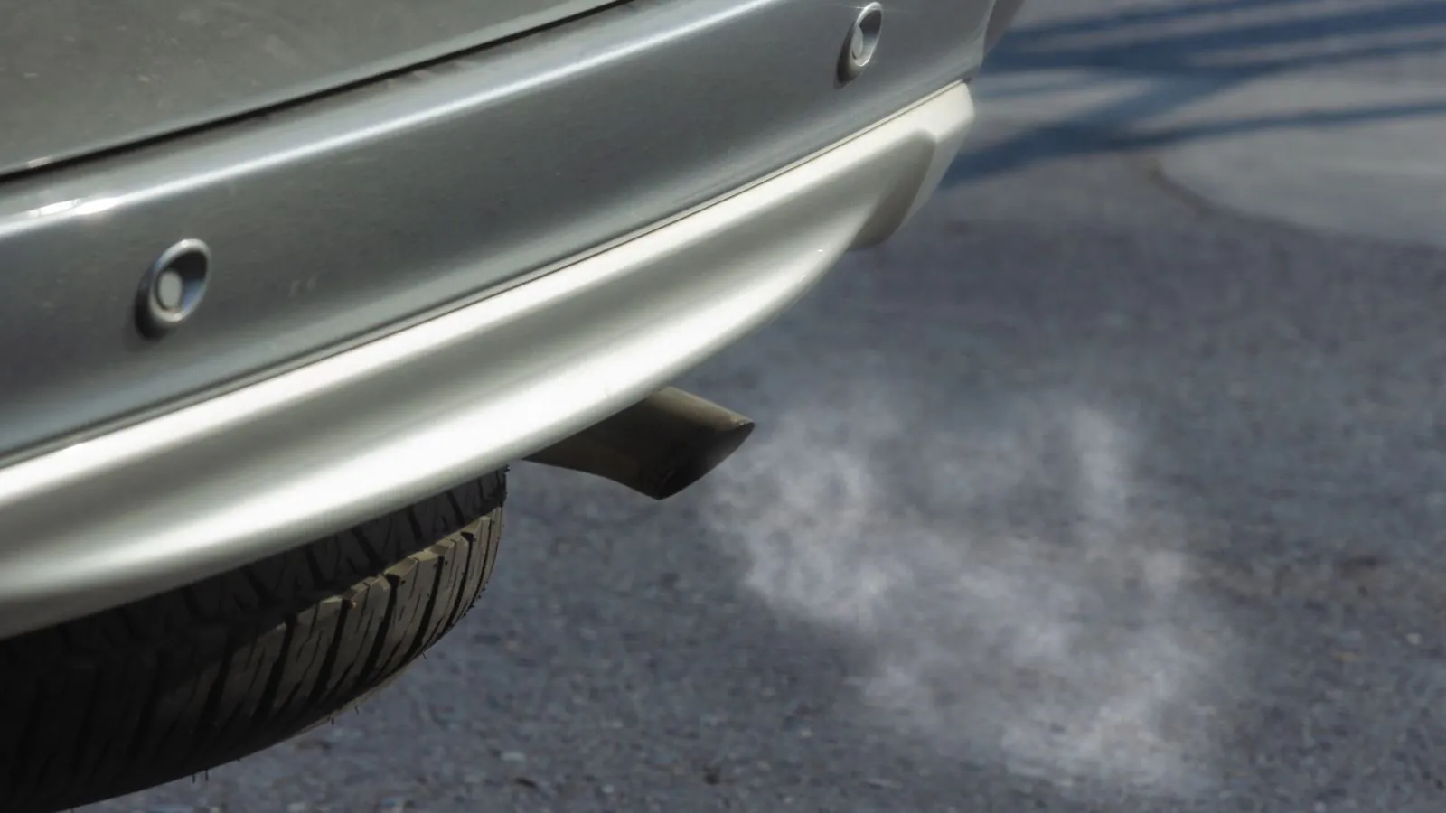 Close-up of a silver car exhaust pipe emitting smoke on asphalt road.