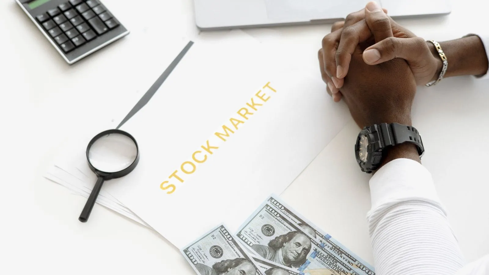Hands folded over stock market documents with calculator and cash on desk.