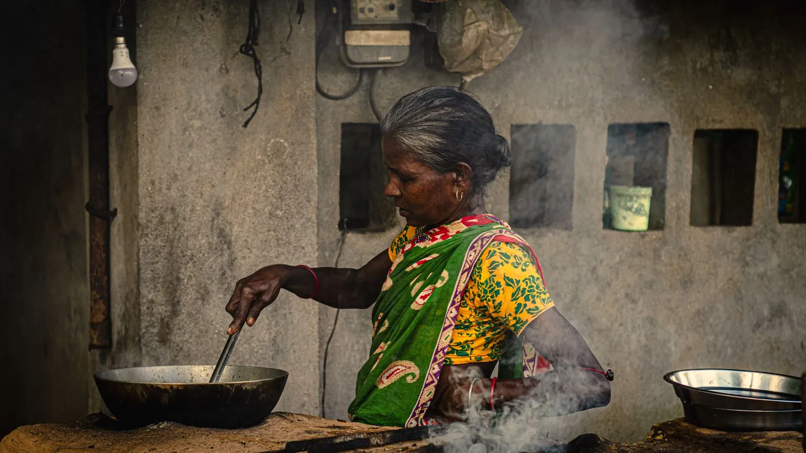 A rural Indian woman in vibrant clothing prepares food outdoors using traditional methods.