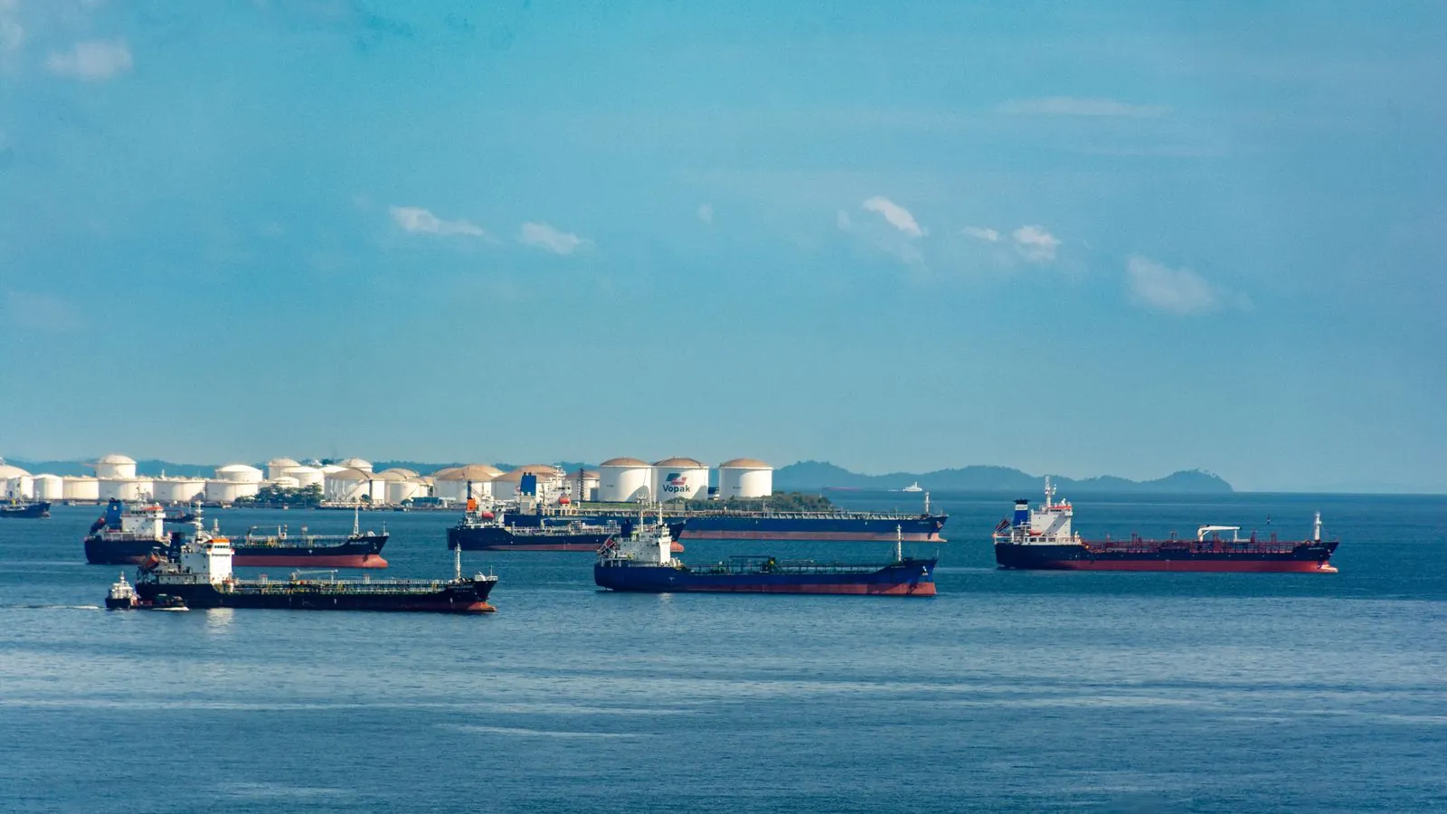 A fleet of cargo ships docked near oil storage tanks along a serene coastline with a clear blue sky above.