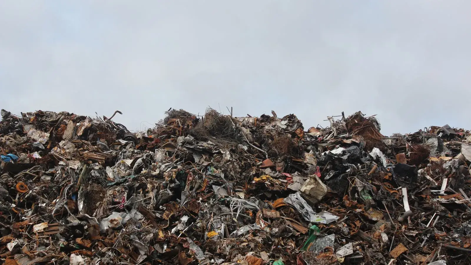 A large pile of scrap metal and waste at a landfill against a cloudy sky.
